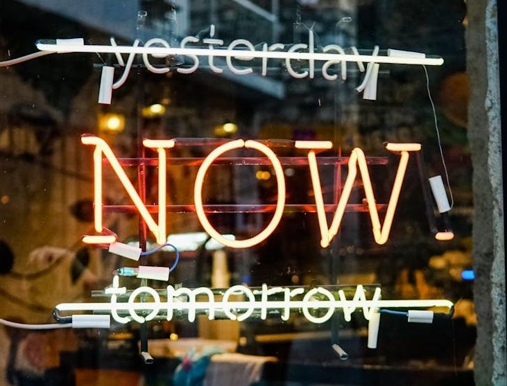 A close-up of a window displaying three neon signs: "yesterday" in white neon at the top, "NOW" in bright orange neon prominently in the middle, and "tomorrow" in yellow-green neon at the bottom. Wires and reflections are visible on the glass.