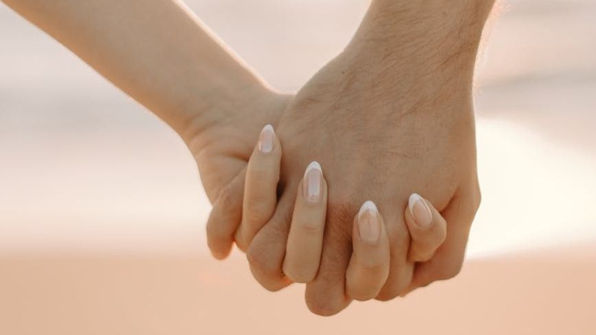 A close-up horizontal shot shows two hands, one appearing to be a man's and the other a woman's, gently clasped together with fingers intertwined. The lighting is soft, suggesting an intimate moment.