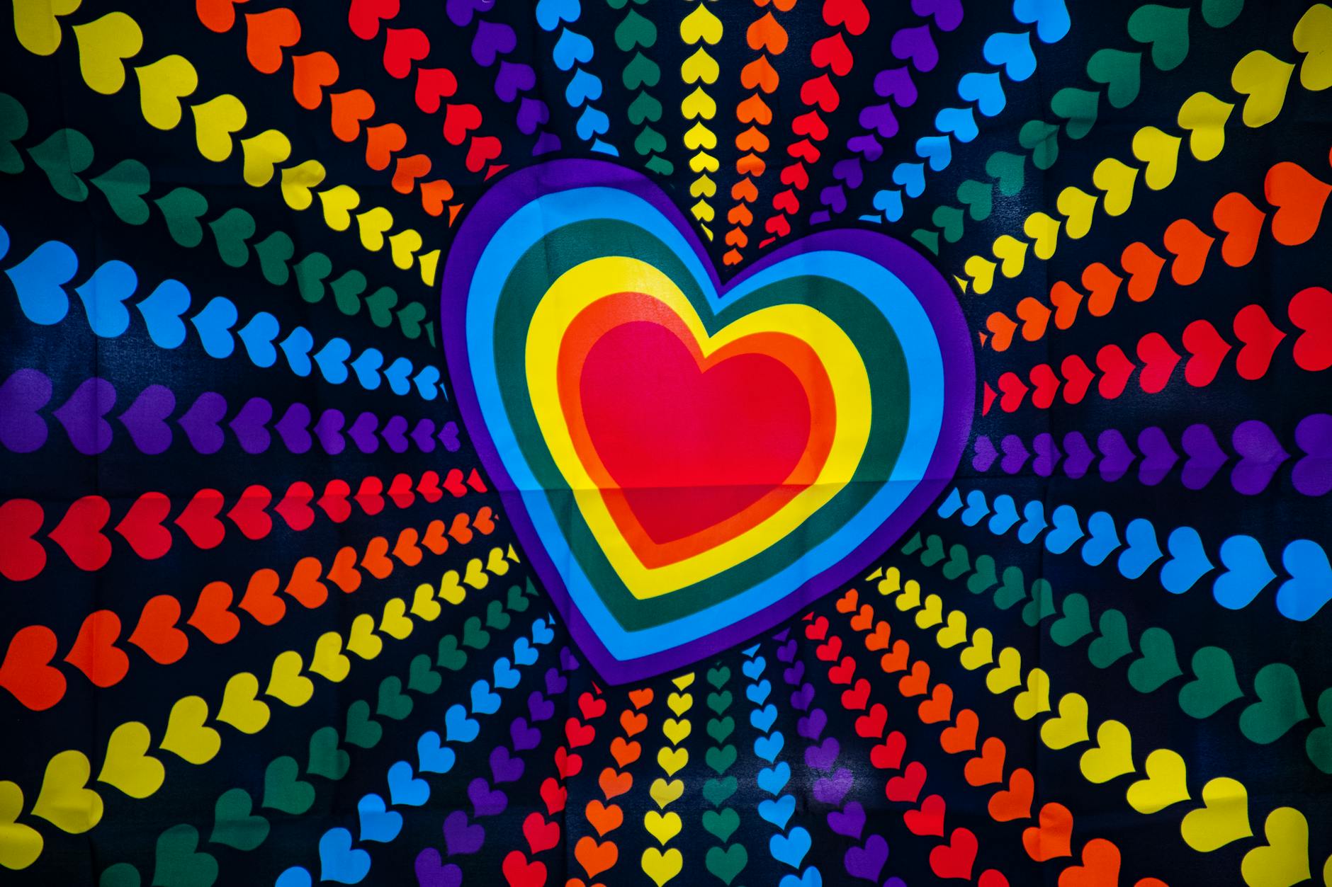 A vibrant, psychedelic pattern featuring a large, multi-colored heart at the center, layered with concentric rainbow stripes of hearts. Smaller, similarly colored hearts radiate outwards from the center in rows, against a dark background, creating a sense of depth and movement.