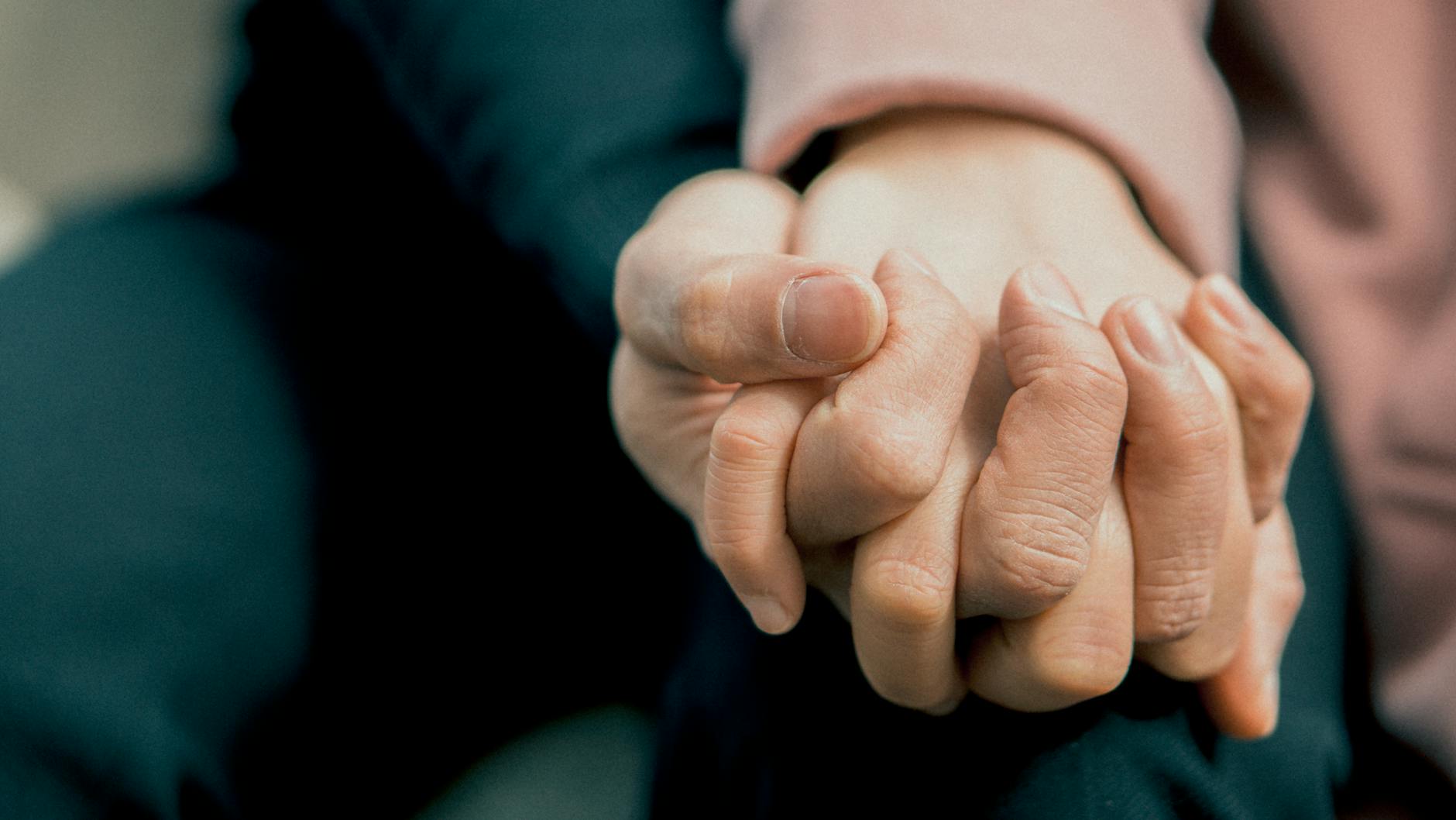 A close-up shot shows two hands clasped together, suggesting a tender or supportive connection. One person wears dark clothing, while the other wears a light pink sleeve.