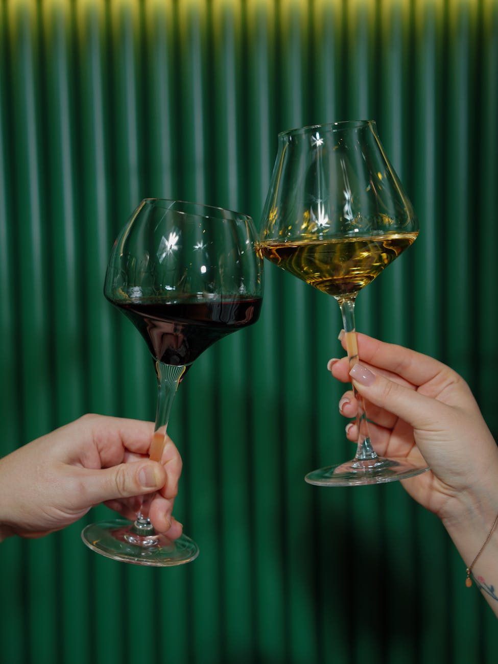 
Two hands hold up elegant wine glasses, one filled with red wine and the other with white wine, against a ribbed, dark green background. The glasses have long, slender stems and uniquely shaped bowls.