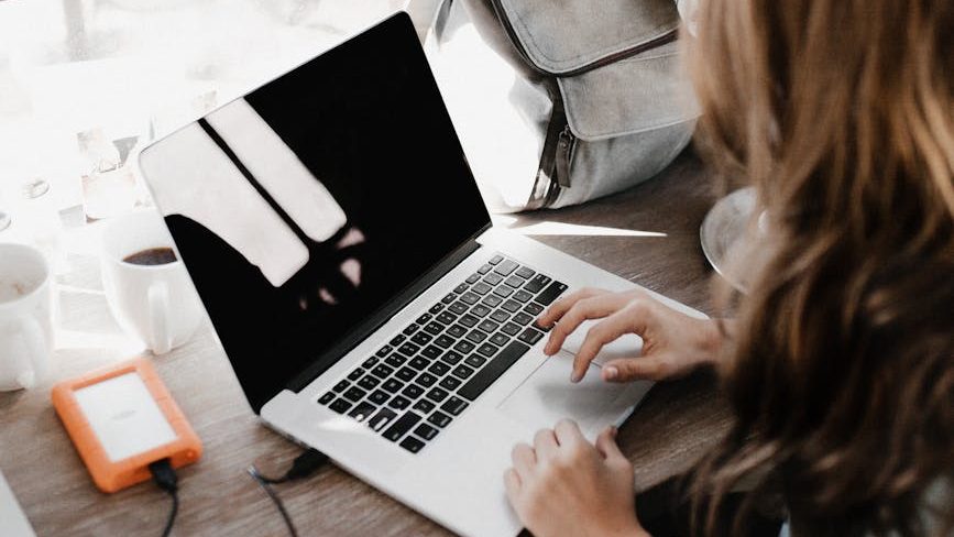 
A person, viewed from over their left shoulder, is typing on a silver laptop. To the left of the laptop are a white mug of coffee and an orange external hard drive. A gray bag is visible in the upper right background.