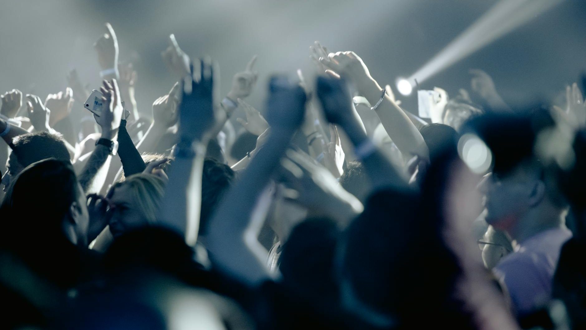 A blurry, low-light photograph captures a crowd of people at what appears to be a concert or event, with many hands raised in the air, silhouetted by bright spotlights from above. This energetic scene embodies the spirit of "The Safety Dance." Some individuals are holding up cell phones.
