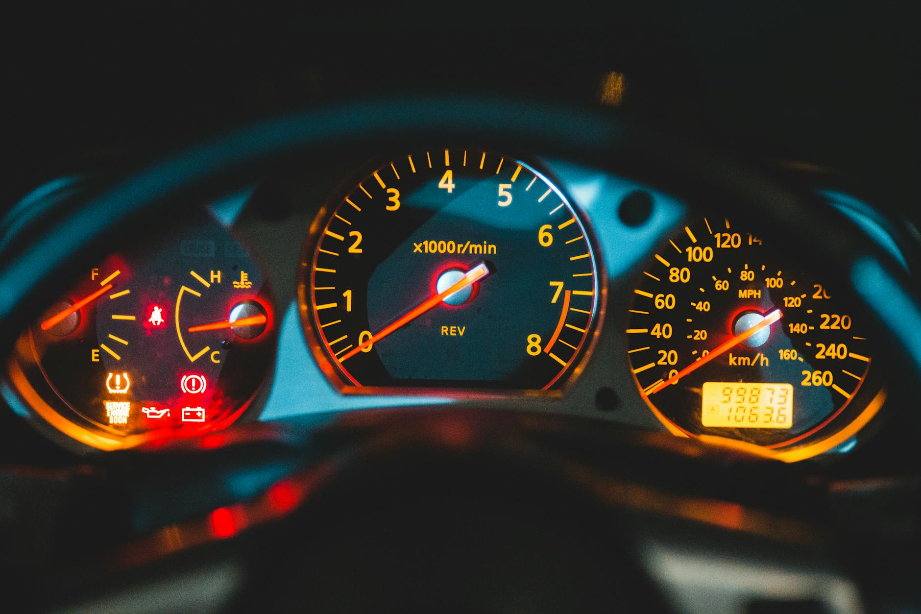 The dashboard of a car at night, illuminated with orange and red lights. Three main gauges are visible: a fuel and temperature gauge on the left with warning lights, a large tachometer in the center, and a speedometer on the right with an odometer. This iconic scene is central to "Paradise by the Dashboard Light."