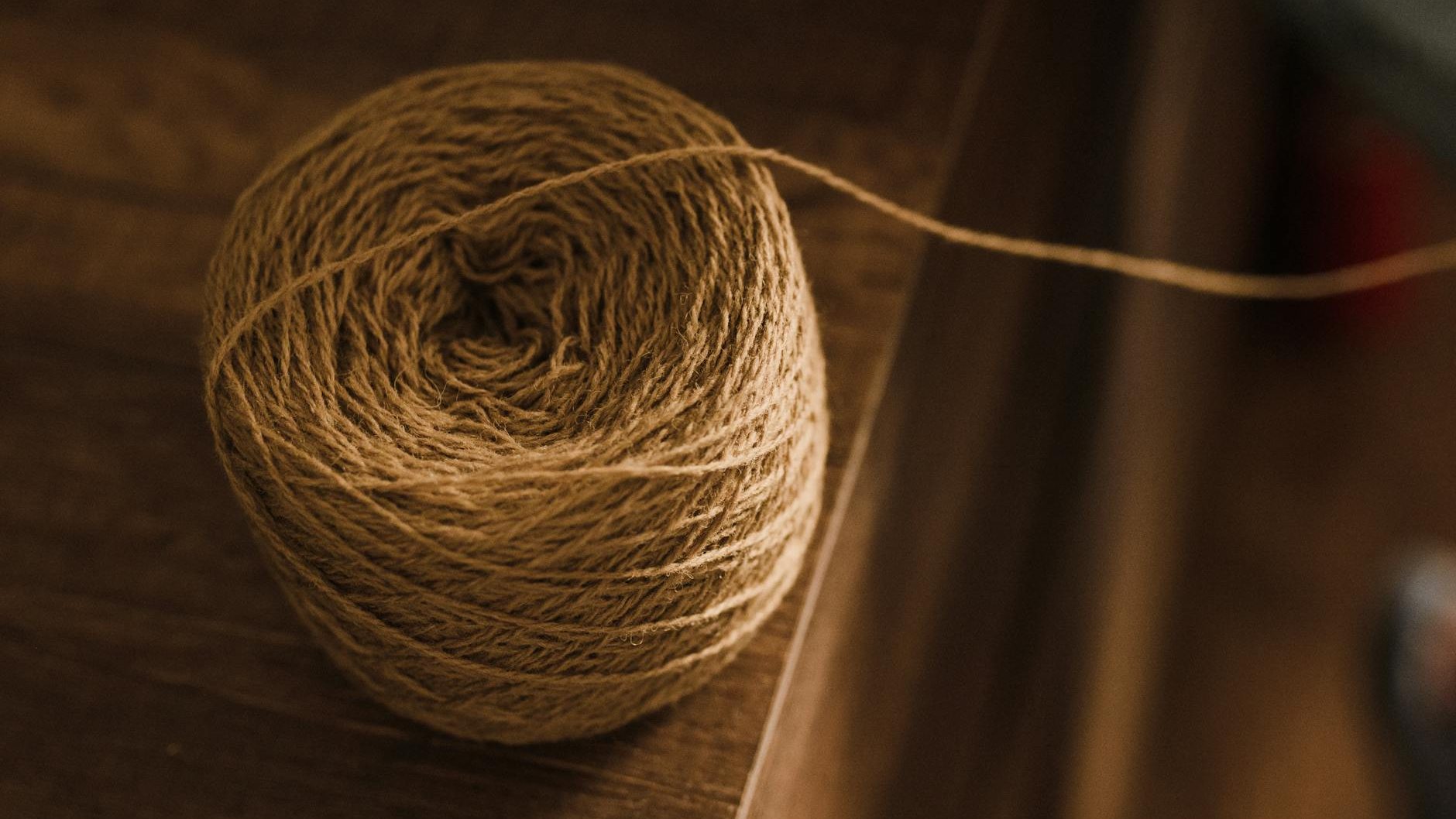 
A neatly wound ball of light brown twine or yarn rests on a wooden surface. A single strand of the material extends from the ball towards the right side of the frame. The background is dimly lit.